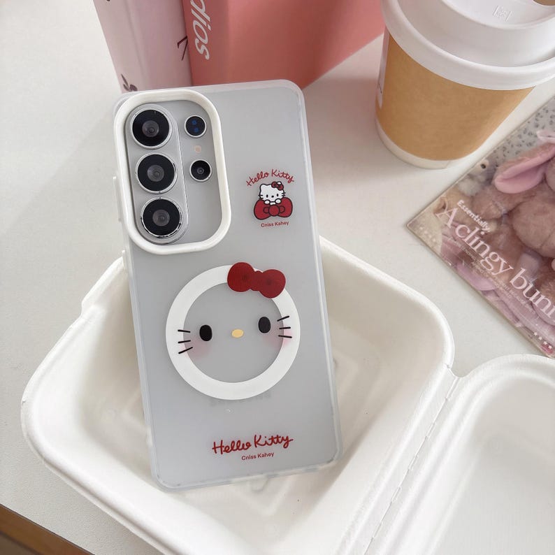 May include: A clear phone case featuring a Hello Kitty design. The case has a gray background with a white border around the camera and a Hello Kitty graphic with a red bow. The words "Hello Kitty" are printed at the bottom.