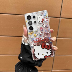 May include: Clear phone case featuring a Hello Kitty design with red glitter and various Hello Kitty character illustrations. The case has a camera cutout and is designed for a smartphone.