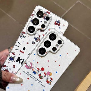K&ouml;nnte beinhalten: Zwei wei&szlig;e Handyh&uuml;llen mit einem Hello Kitty-Design. Die H&uuml;llen zeigen ein sich wiederholendes Muster der Figur, zusammen mit dem Text "Ki" und "Let's go on adventures together, play together, and keep each other company."