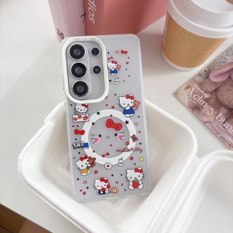 May include: Clear phone case featuring a Hello Kitty design. The case has a gray background with multiple Hello Kitty illustrations, bows, and other decorative elements. The text "Hello Kitty" is visible on the case. The case is designed to fit a phone with a triple-lens camera.