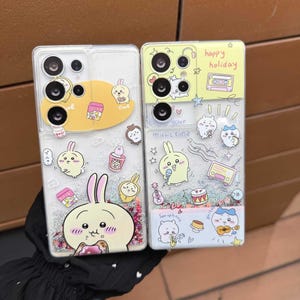 以下が含まれることがあります： カラフルな漫画のデザインが施された、2つの透明な携帯電話ケース。1つのケースには「time」の文字が入った黄色のウサギが描かれており、もう1つには「happy holiday」と「your music time」の文字が入っています。どちらのケースにもキラキラが付いています。
