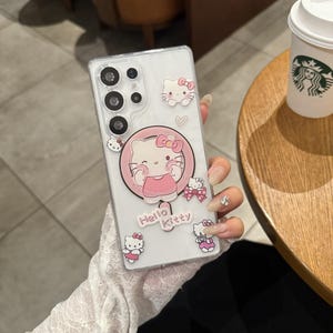 以下が含まれることがあります： ハローキティのデザインが施された透明な携帯電話ケース。ケースには、ピンクの円の中に大きなハローキティのグラフィックがあり、端には小さなハローキティのステッカーが付いています。「Hello Kitty」の文字が下部に印刷されています。携帯電話ケースは人が持っています。