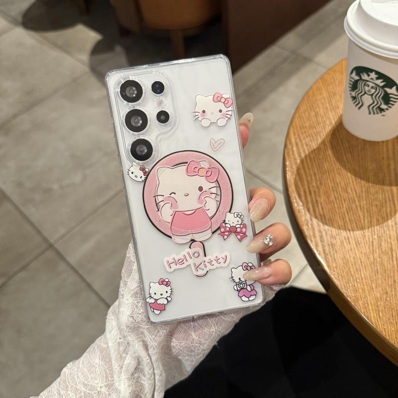 以下が含まれることがあります： ハローキティのデザインが施された透明な携帯電話ケース。ケースには、ピンクの円の中に大きなハローキティのグラフィックがあり、端には小さなハローキティのステッカーが付いています。「Hello Kitty」の文字が下部に印刷されています。携帯電話ケースは人が持っています。