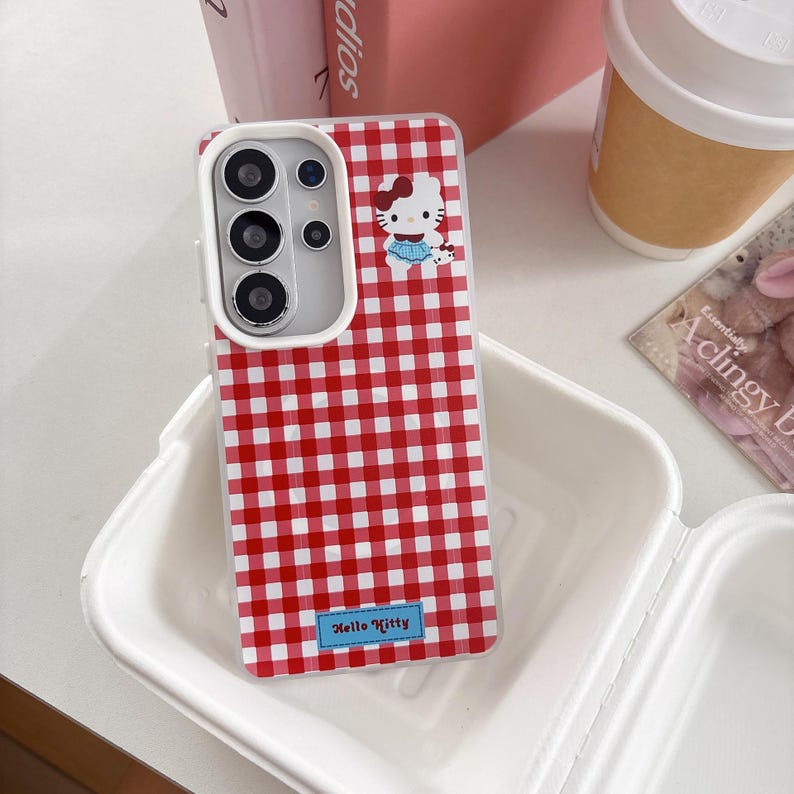 May include: A red and white checkered phone case featuring a Hello Kitty design. The case has a white border and a small blue rectangle with the words "Hello Kitty". The phone case is in a white container.