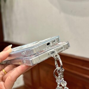 Może przedstawiać: Dwa przezroczyste etui na telefon ułożone jedno na drugim. Etui mają kwiatowy wz&oacute;r i srebrny metalowy pierścień przymocowany do koralikowego uroku. Etui są wykonane z przezroczystego materiału.