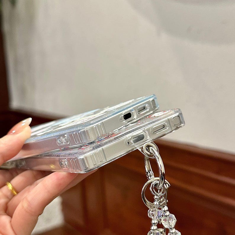 Może przedstawiać: Dwa przezroczyste etui na telefon ułożone jedno na drugim. Etui mają kwiatowy wz&oacute;r i srebrny metalowy pierścień przymocowany do koralikowego uroku. Etui są wykonane z przezroczystego materiału.