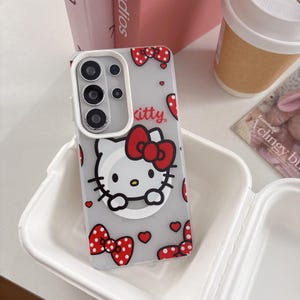 May include: A clear phone case featuring a Hello Kitty design with a red bow and red polka dot bows. The case has a gray background with red hearts and the word "kitty". The phone case has a white border.