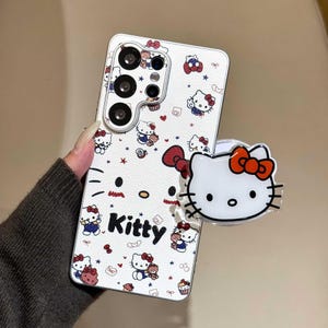 K&ouml;nnte beinhalten: Wei&szlig;es Handycase mit einem sich wiederholenden Muster von Hello Kitty-Figuren, roten Schleifen und Sternen. Das Wort "Kitty" ist in Schwarz gedruckt. Ein passender wei&szlig;er Pop-Socket mit einem Hello Kitty-Gesicht und einer roten Schleife ist angebracht.