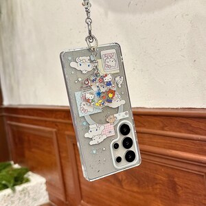 Może przedstawiać: Przezroczyste etui na telefon z kresk&oacute;wkowym wzorem z Hello Kitty i innymi postaciami. Etui ma srebrny łańcuszek i ozdobny urok. Projekt zawiera chmury, gwiazdy i r&oacute;żne kolorowe ilustracje.