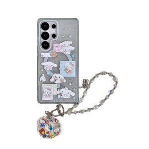 Może przedstawiać: Przezroczyste etui na telefon z kresk&oacute;wkowym wzorem z Hello Kitty i innymi postaciami. Etui ma srebrny łańcuszek z perłami i zawieszkę z postaciami z kresk&oacute;wek. Etui na telefon jest przeznaczone do smartfona.