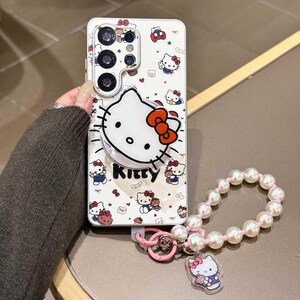 K&ouml;nnte beinhalten: Wei&szlig;es Handycase mit Hello Kitty-Design und Pop-up-Griff. Das Case hat ein sich wiederholendes Muster von Hello Kitty-Charakteren und Schleifen. Eine Perlen-Handgelenksschlaufe mit Hello Kitty-Anh&auml;nger ist befestigt.