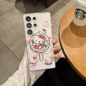 以下が含まれることがあります： ピンクと白のハローキティのデザインで装飾された透明な携帯電話ケース。ケースには、複数のハローキティの画像、ピンクのリボン、そして「Have a good day」という言葉が特徴です。