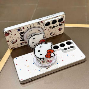 K&ouml;nnte beinhalten: Wei&szlig;es Handycase mit silbernem Rahmen, mit einem sich wiederholenden Muster von Hello Kitty-Charakteren und dem Wort "Kitty". Ein klarer Handygriff mit Hello Kitty-Design ist angebracht. Das Case hat einen Kameraausschnitt und bunte Punkte.