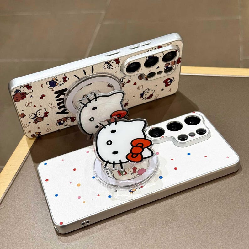 K&ouml;nnte beinhalten: Wei&szlig;es Handycase mit silbernem Rahmen, mit einem sich wiederholenden Muster von Hello Kitty-Charakteren und dem Wort "Kitty". Ein klarer Handygriff mit Hello Kitty-Design ist angebracht. Das Case hat einen Kameraausschnitt und bunte Punkte.