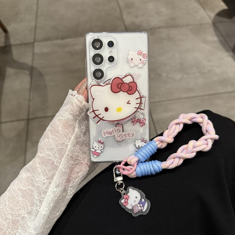 以下が含まれることがあります： ハローキティのグラフィックが施された透明な携帯電話ケース。ケースにはピンクのリボン、「Hello Kitty」の文字、ピンクとブルーの編み込みリストストラップが付いています。ハローキティのデザインの小さなチャームが取り付けられています。