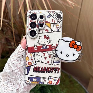 Peut inclure: Coque de t&eacute;l&eacute;phone blanche avec un motif Hello Kitty color&eacute;, avec des touches de rouge, de bleu et de jaune. La coque est orn&eacute;e d'un support pop-up avec le visage de Hello Kitty et un n&oelig;ud orange. Le texte "HELLO KITTY" est imprim&eacute;.