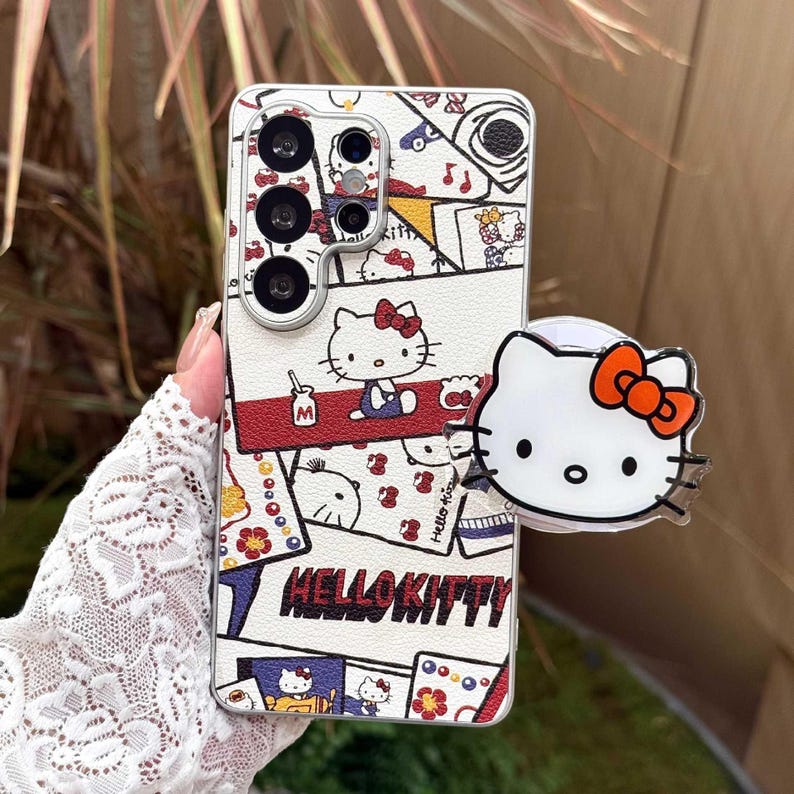 Peut inclure: Coque de t&eacute;l&eacute;phone blanche avec un motif Hello Kitty color&eacute;, avec des touches de rouge, de bleu et de jaune. La coque est orn&eacute;e d'un support pop-up avec le visage de Hello Kitty et un n&oelig;ud orange. Le texte "HELLO KITTY" est imprim&eacute;.