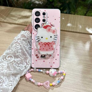 Peut inclure: Coque de t&eacute;l&eacute;phone rose avec un motif Hello Kitty, accompagn&eacute;e d'un bracelet assorti. L'&eacute;tui pr&eacute;sente un motif d'&eacute;toiles et une figurine Hello Kitty en 3D. Le bracelet comprend des perles roses, bleues et transparentes, avec un charm Hello Kitty et un charm c&oelig;ur rose.