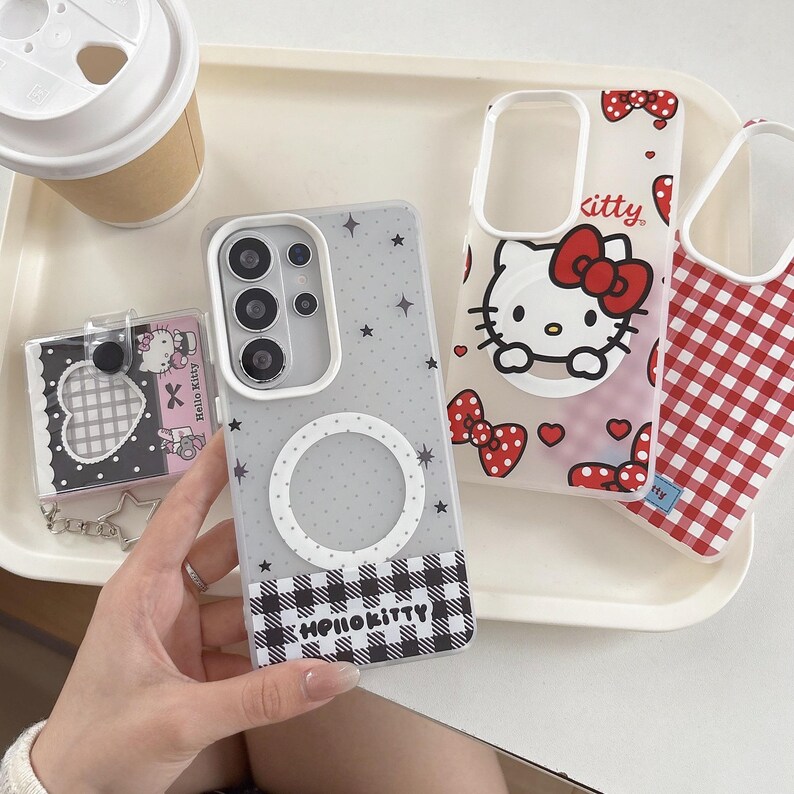 May include: A collection of Hello Kitty phone cases and accessories. One case is clear with a black and white gingham pattern and the text "Hello Kitty". Another case features the Hello Kitty character with a red bow. A small card holder and a coffee cup are also visible.