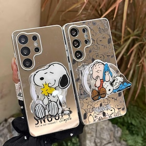 Könnte beinhalten: Zwei verspiegelte Smartphone-Hüllen mit Peanuts-Figuren. Die linke Hülle zeigt Snoopy und Woodstock mit dem Text "SNOOPY You're not relaxed! Simple". Die rechte Hülle hat ein wiederkehrendes Snoopy-Muster und einen Linus- und Snoopy-Halter.
