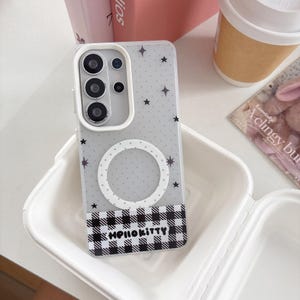May include: A clear phone case with a gray background, featuring a pattern of black stars and a white circle. The bottom section has a black and white checkered pattern with the text "Hello Kitty". The case is in a white container.