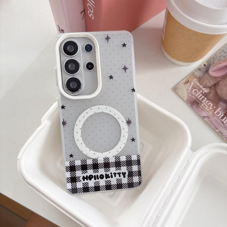 May include: A clear phone case with a gray background, featuring a pattern of black stars and a white circle. The bottom section has a black and white checkered pattern with the text "Hello Kitty". The case is in a white container.