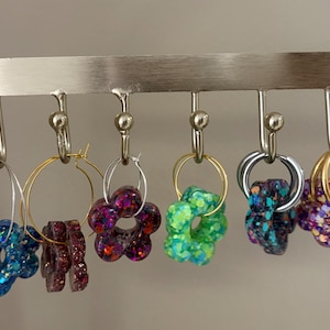 May include: A display rack holds six pairs of earrings. The earrings feature various designs, including flower shapes and hoops, with colors like blue, purple, green, and gold. The earrings are adorned with glitter.