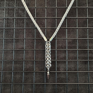 Könnte beinhalten: Eine silberfarbene Halskette mit einer geflochtenen Kette, die an einen zentralen vertikalen Anhänger anschließt. Der Anhänger besteht aus ineinandergreifenden Metallringen in Kettenpanzeroptik und endet in einem kleinen, spitzen Metallkegel.