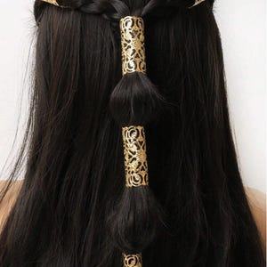 Puede incluir: Primer plano de cabello largo y oscuro peinado con una trenza y puños dorados decorativos. Los puños son cilíndricos con intrincados diseños calados. Los accesorios para el cabello añaden un toque de elegancia.