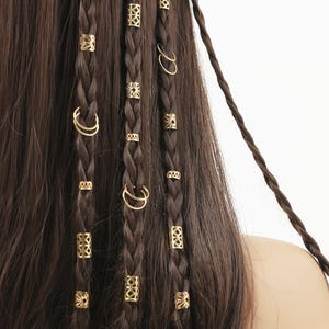 Puede incluir: Primer plano de cabello castaño trenzado adornado con puños y anillos dorados. Los accesorios para el cabello presentan varios diseños geométricos y florales. Las trenzas son de color marrón oscuro, con un fondo liso.