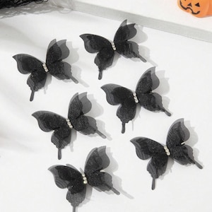 May include: Seven black butterfly decorations with rhinestone accents. The butterflies have a sheer, layered wing design and are arranged on a white surface. A small orange pumpkin decoration is in the background.