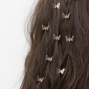 Può includere: Primo piano di capelli castani adornati con piccole mollette dorate a forma di farfalla. Le mollette sono uniformemente distanziate tra i capelli mossi, aggiungendo un tocco delicato e stravagante all'acconciatura. Lo sfondo è bianco.