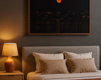 May include: A bedroom scene featuring a large framed print of a full moon over a city at night. The bed has neutral-toned bedding with brown pillows and a rust-colored throw. A wooden nightstand holds a lamp.