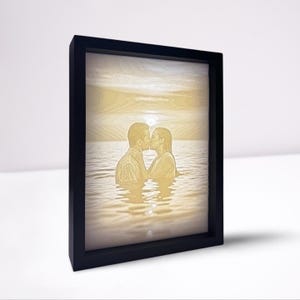 Könnte beinhalten: Ein gerahmtes, lasergraviertes Kunstwerk, das ein sich küssendes Paar im Meer bei Sonnenuntergang zeigt. Das Bild ist in Gelb- und Brauntönen gehalten und in einem schwarzen Rahmen eingefasst. Das Kunstwerk fängt einen romantischen Moment ein.