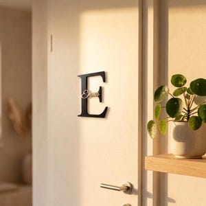 Könnte beinhalten: Ein schwarzer Metallbuchstabe "E" an einer weißen Tür. Der Buchstabe hat einen kursiven Schriftzug "Emma". Die Tür hat einen silbernen Griff und ein Schlüsselloch. Eine kleine Topfpflanze steht auf einem Holzregal.