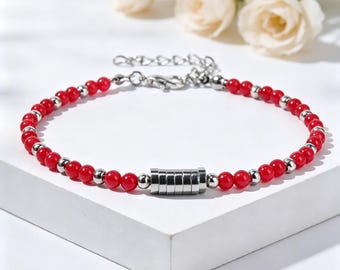 May include: A beaded bracelet featuring alternating small red spherical beads and tiny silver-toned metallic spheres. The center of the bracelet displays a cylindrical silver-toned metal component composed of several stacked rings. It includes a silver-toned chain extender and a lobster claw clasp.