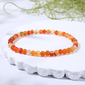 May include: A circular beaded bracelet featuring small, polished, spherical stones in varying shades of orange, red, and translucent amber. The beads are strung together in a continuous loop, displaying natural color gradients and subtle internal textures.