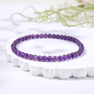 May include: A circular bracelet composed of small, smooth, polished purple amethyst beads. The beads are uniform in size and strung together in a continuous loop, showcasing the natural translucent violet hues and subtle variations of the gemstone.