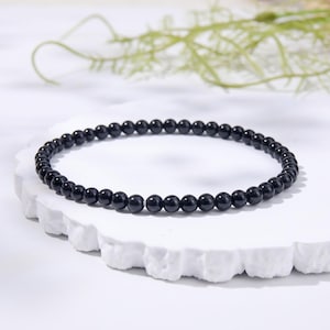 Puede incluir: Una pulsera circular compuesta por pequeñas cuentas esféricas negras, uniformes y pulidas. Las cuentas están ensartadas en un bucle continuo, creando un diseño sencillo. Las cuentas negras tienen un acabado de superficie liso y brillante.