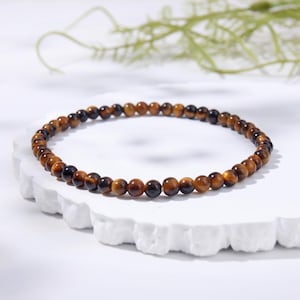 Puede incluir: Una pulsera circular compuesta por pequeñas cuentas pulidas de ojo de tigre. Las cuentas presentan un patrón natural de bandas con tonos alternos de marrón dorado, ámbar y carbón oscuro, creando una superficie lisa y reflectante en toda la pieza.