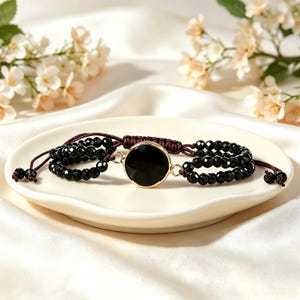 Può includere: Un bracciale con una pietra nera circolare centrale incastonata in una cornice di metallo dorato, affiancata da due file di perline nere sfaccettate su ciascun lato. Il gioiello è realizzato con un cordino marrone scuro e un nodo scorrevole.