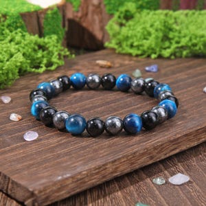 May include: A beaded bracelet featuring a repeating pattern of polished round beads in vibrant blue, glossy black, and metallic grey. The beads are strung together in a circular arrangement, showcasing a smooth, reflective surface on each individual stone.