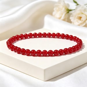 Puede incluir: Una pulsera roja de cuentas compuesta por pequeñas cuentas redondas. La pulsera se muestra sobre una superficie blanca con un fondo blanco suave y algunas flores blancas.