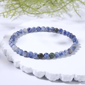 May include: A beaded bracelet composed of small, round, polished stones in varying shades of blue, white, and grey, featuring a single dark grey bead and a single green bead interspersed among the blue and white marbled patterned beads.