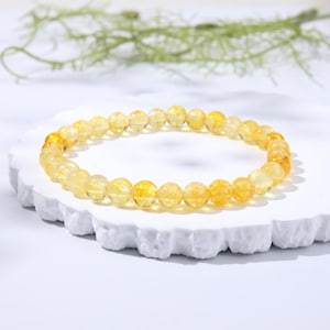 May include: A circular bracelet composed of numerous small, polished, translucent yellow beads. Each bead features internal golden inclusions and a smooth, reflective surface. The beads are strung together in a continuous loop, creating a uniform, beaded design.