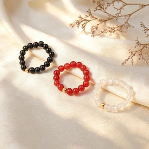 May include: Three circular rings made of small, smooth beads. One ring features black beads, one features red beads, and one features translucent white beads. Each ring includes a single, small, polished gold-tone metal bead accent.