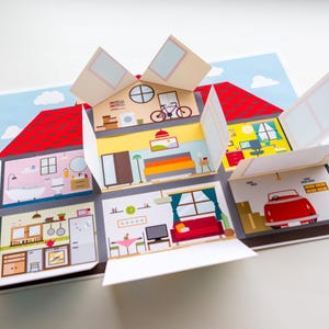 Könnte beinhalten: Ein aufklappbares Papierhaus mit rotem Dach und mehreren Zimmern, darunter ein Badezimmer, ein Schlafzimmer, eine Küche und eine Garage. Jedes Zimmer ist mit Möbeln und Accessoires eingerichtet, vor einem blauen Himmel.