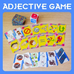 Può includere: Un colorato gioco educativo con il testo "ADJECTIVE GAME". Il gioco include carte con illustrazioni e testo, un dado e una pila di carte con un design a motivi. Il gioco è progettato per insegnare gli aggettivi.