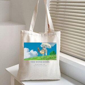 May include: Cream-colored tote bag featuring a colorful illustration from "The Wind Rises." The image shows two figures under an umbrella on a grassy hill with a blue sky. Text at the bottom reads "THE WIND RISES STUDIO GHIBLI 2013".