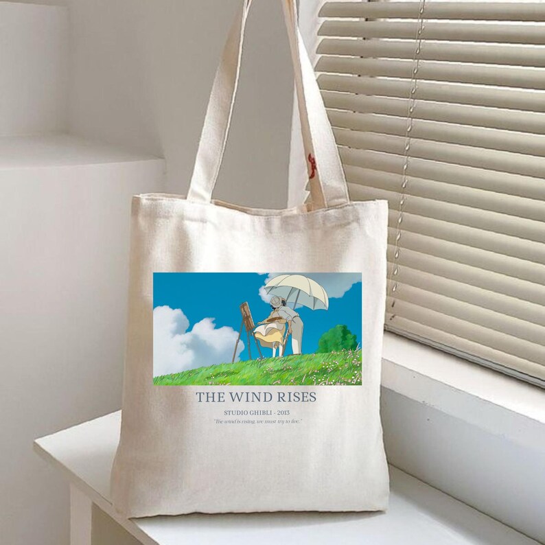 May include: Cream-colored tote bag featuring a colorful illustration from "The Wind Rises." The image shows two figures under an umbrella on a grassy hill with a blue sky. Text at the bottom reads "THE WIND RISES STUDIO GHIBLI 2013".